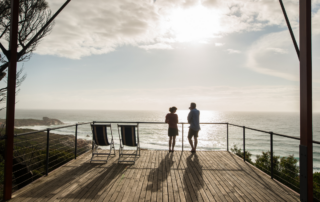 Two guests stand on a wooden deck with modern metal railings, enjoying panoramic views of a rugged