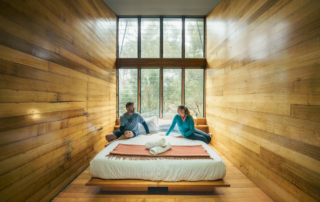 Two guests sit on a bed in a minimalist wooden cabin bedroom overlooking forested landscape through