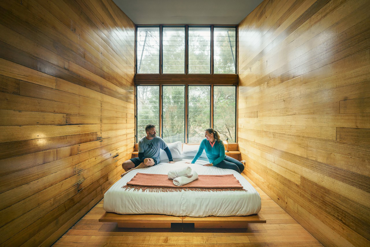 Two guests sit on a bed in a minimalist wooden cabin bedroom overlooking forested landscape through