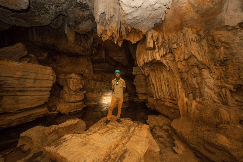 A caver in tan clothing and blue headlamp stands in a spacious underground chamber within Mimbi