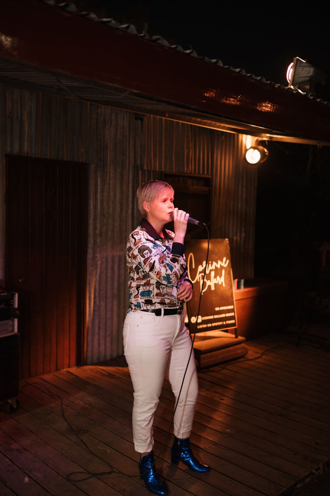A performer with short blonde hair sings into a microphone on a wooden stage, wearing a patterned