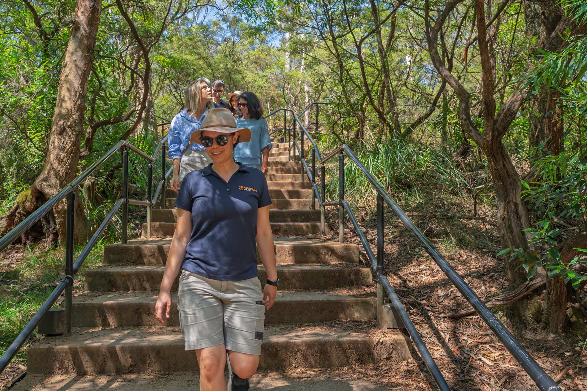 A group of tourists hiking down a scenic wooden staircase through a lush, tree-lined forest path