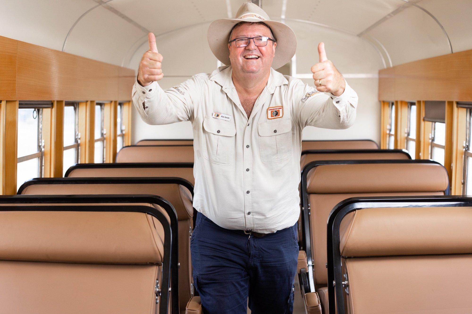 Friendly Tour Guide in Coach Bus Interior A cheerful tour guide wearing a cream-colored shirt, cowboy hat, and glasses gives two thumbs up