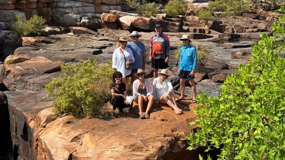 A diverse group of seven hikers poses together on a large, flat rocky outcrop surrounded