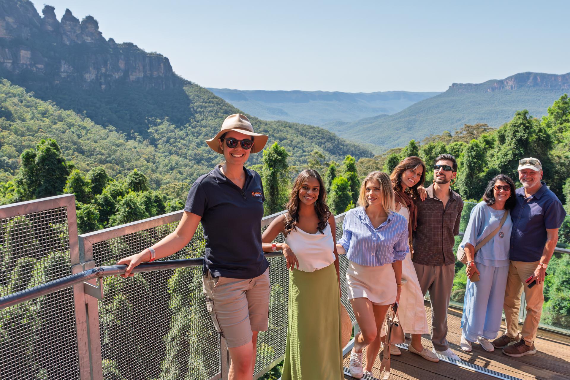 A cheerful group of eight tourists poses together on a scenic lookout platform overlooking