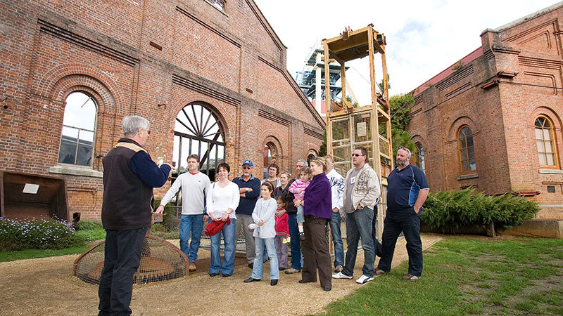 A group of visitors gathered in a courtyard surrounded by heritage brick buildings listen to