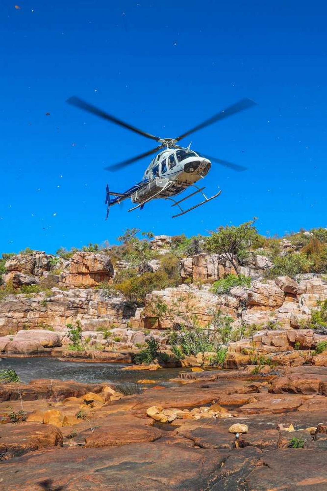 A white and gold helicopter hovers above a rocky riverbed in a remote gorge surrounded by red