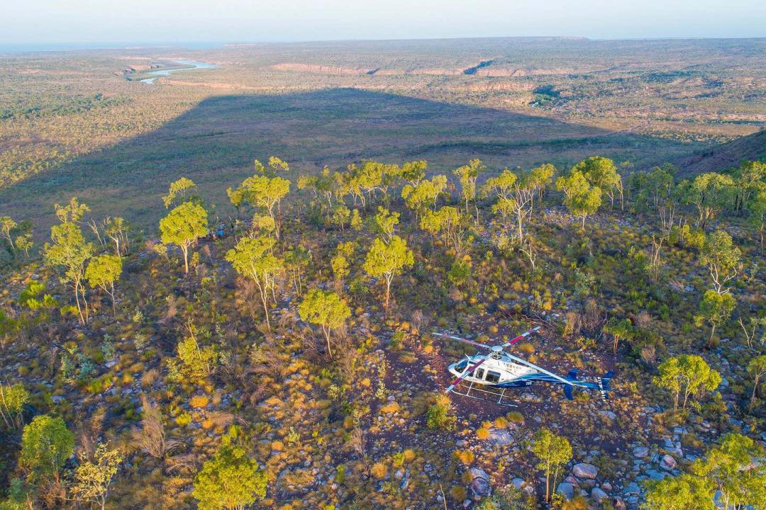 An aerial view of a blue and white helicopter flying over the rugged Australian outback terrain