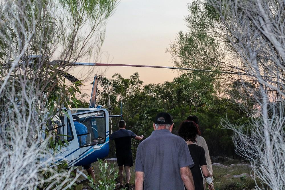A group of tourists prepare to board a blue and white helicopter in a remote, scrubby landscape