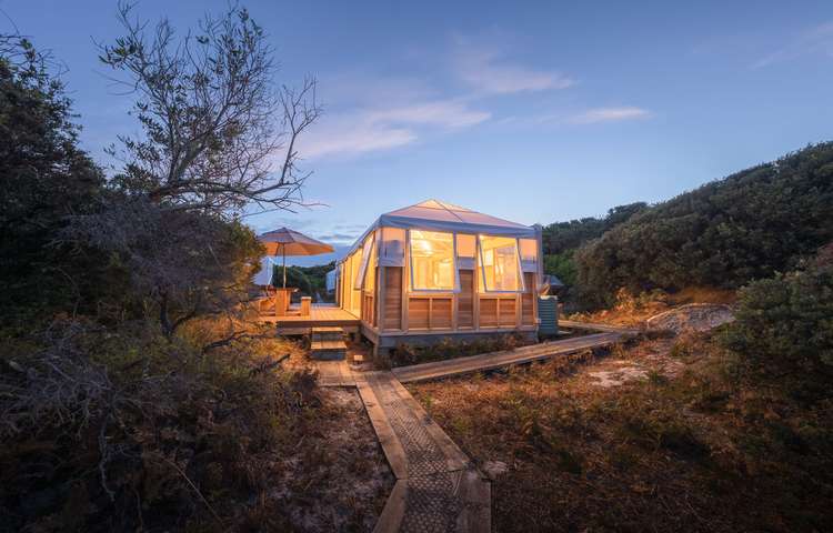 A glamping-style dome cabin glowing at dusk, surrounded by coastal scrub and connected by a timber boardwalk