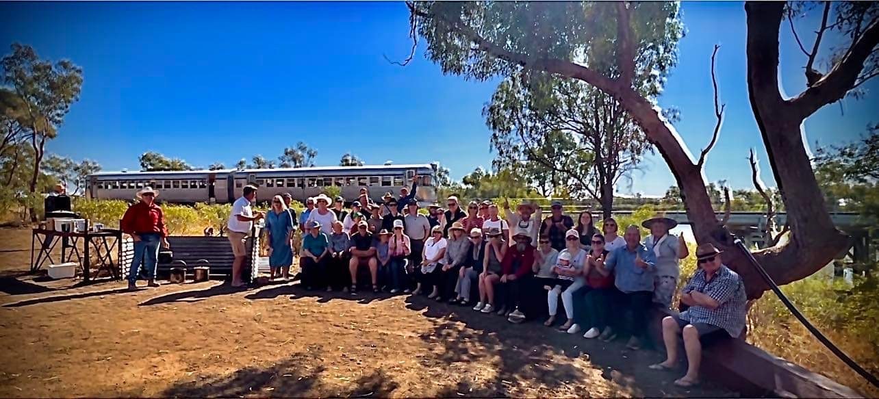 A large group of tourists gathered in the shade of massive eucalyptus trees beside a heritage