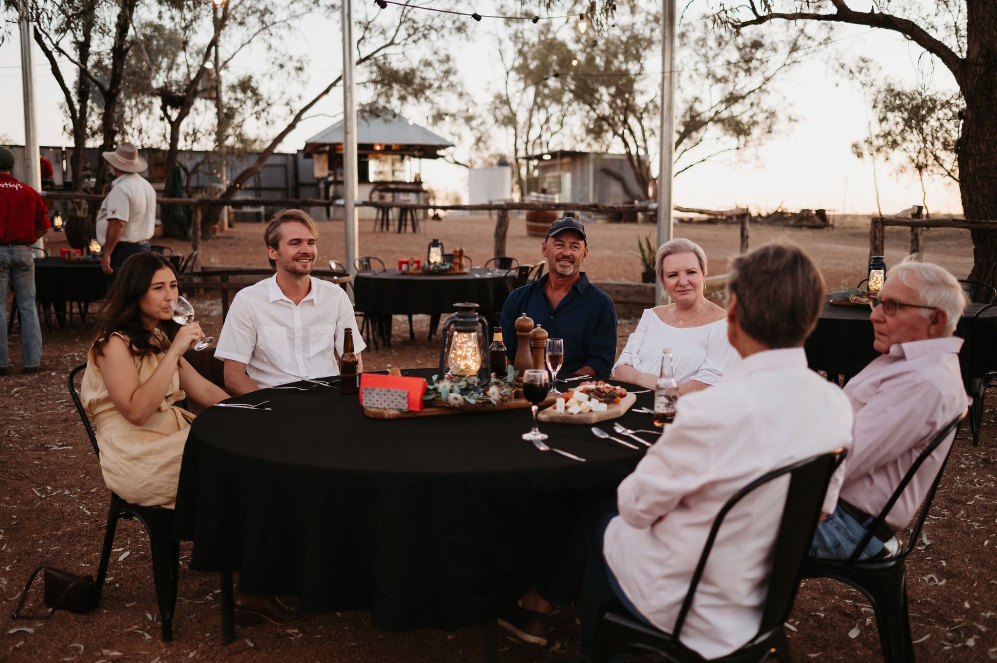 A group of six guests enjoys an elegant dinner gathering under string lights in a remote Australian