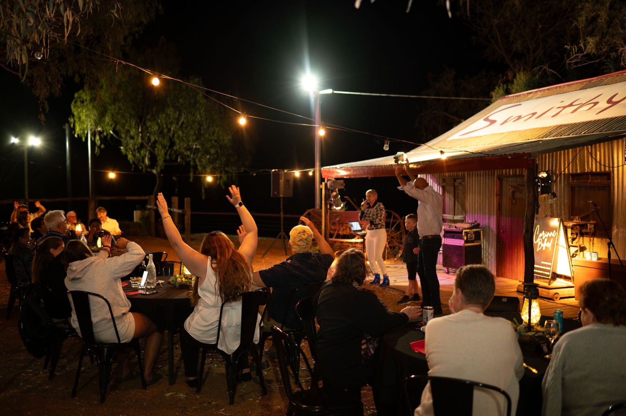 A lively outdoor evening gathering under warm ambient lighting, featuring guests seated at tables