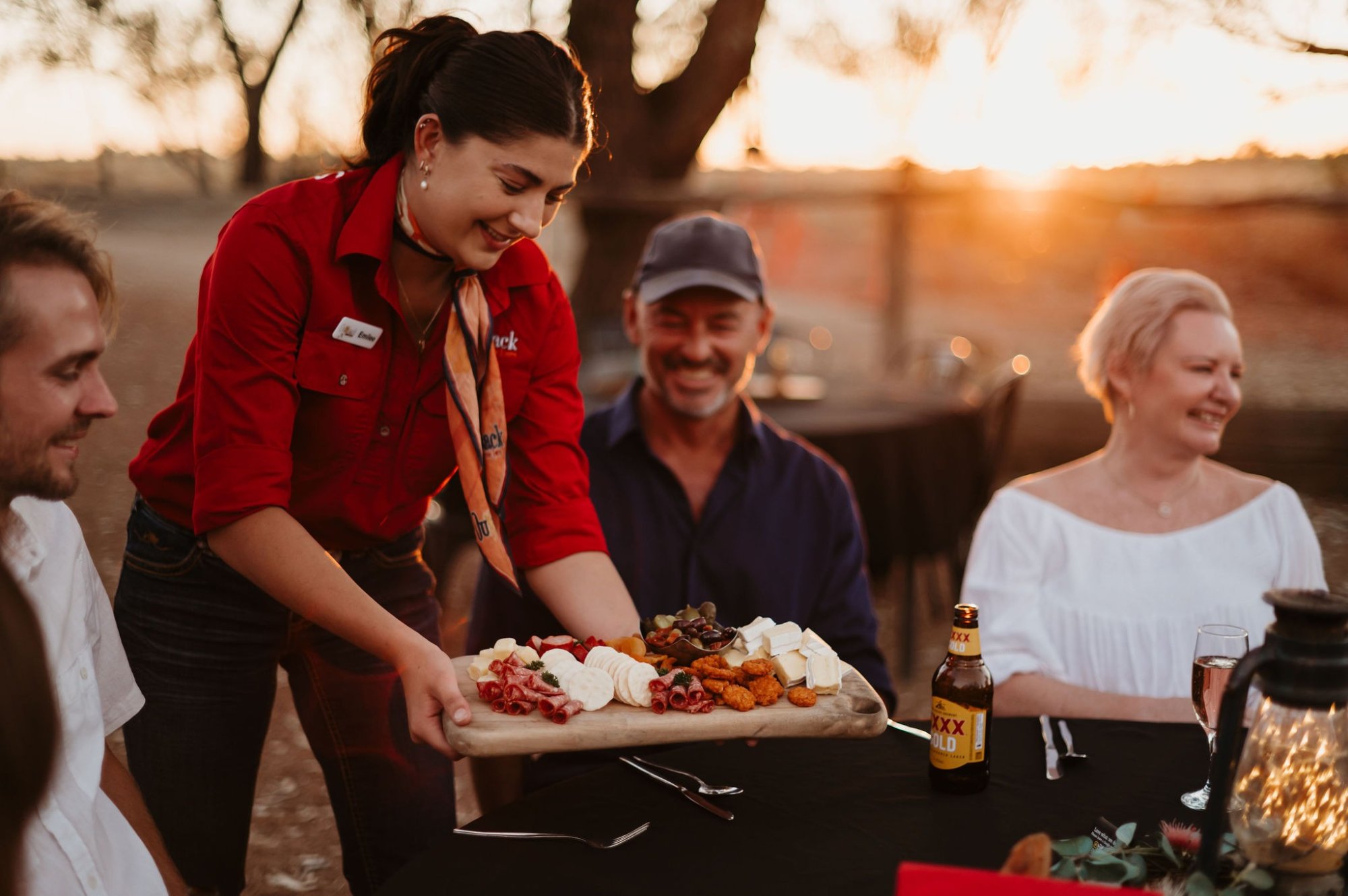 A smiling server in a red uniform presents a wooden board of cured meats and cheese to guests