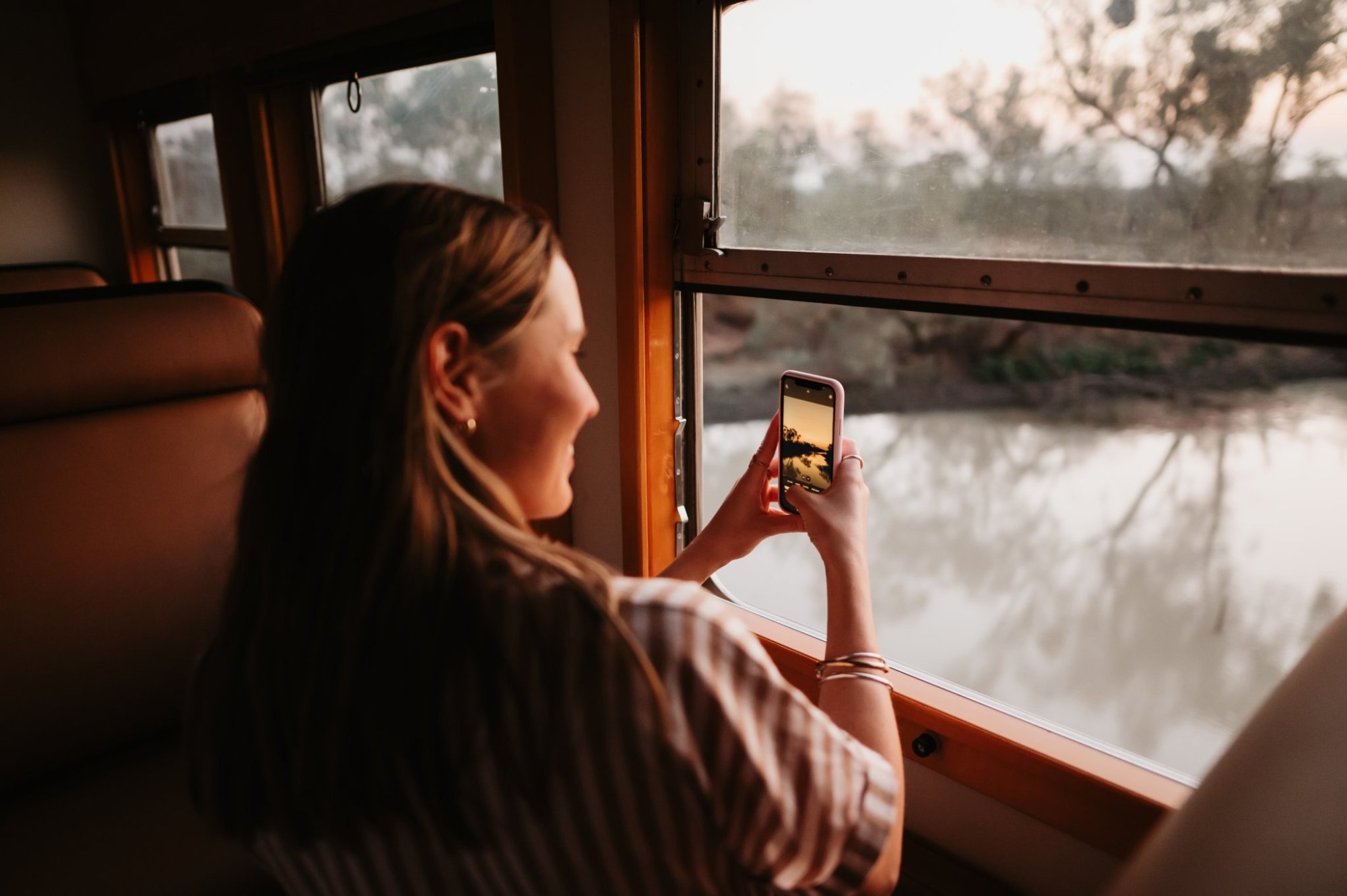 Passenger Photographing River Landscape from Cruise Window A woman stands at a large window inside what appears to be a river cruise vessel, holding up her