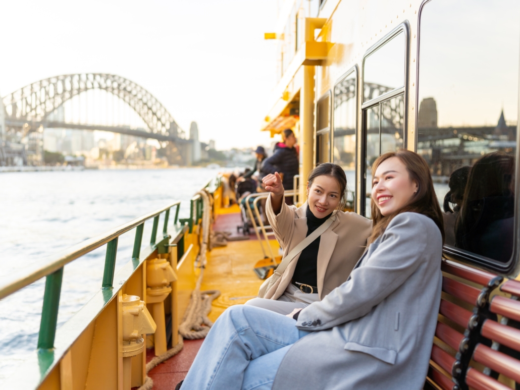 Two young women enjoy the deck of a Sydney Harbour ferry, smiling and pointing toward the iconic