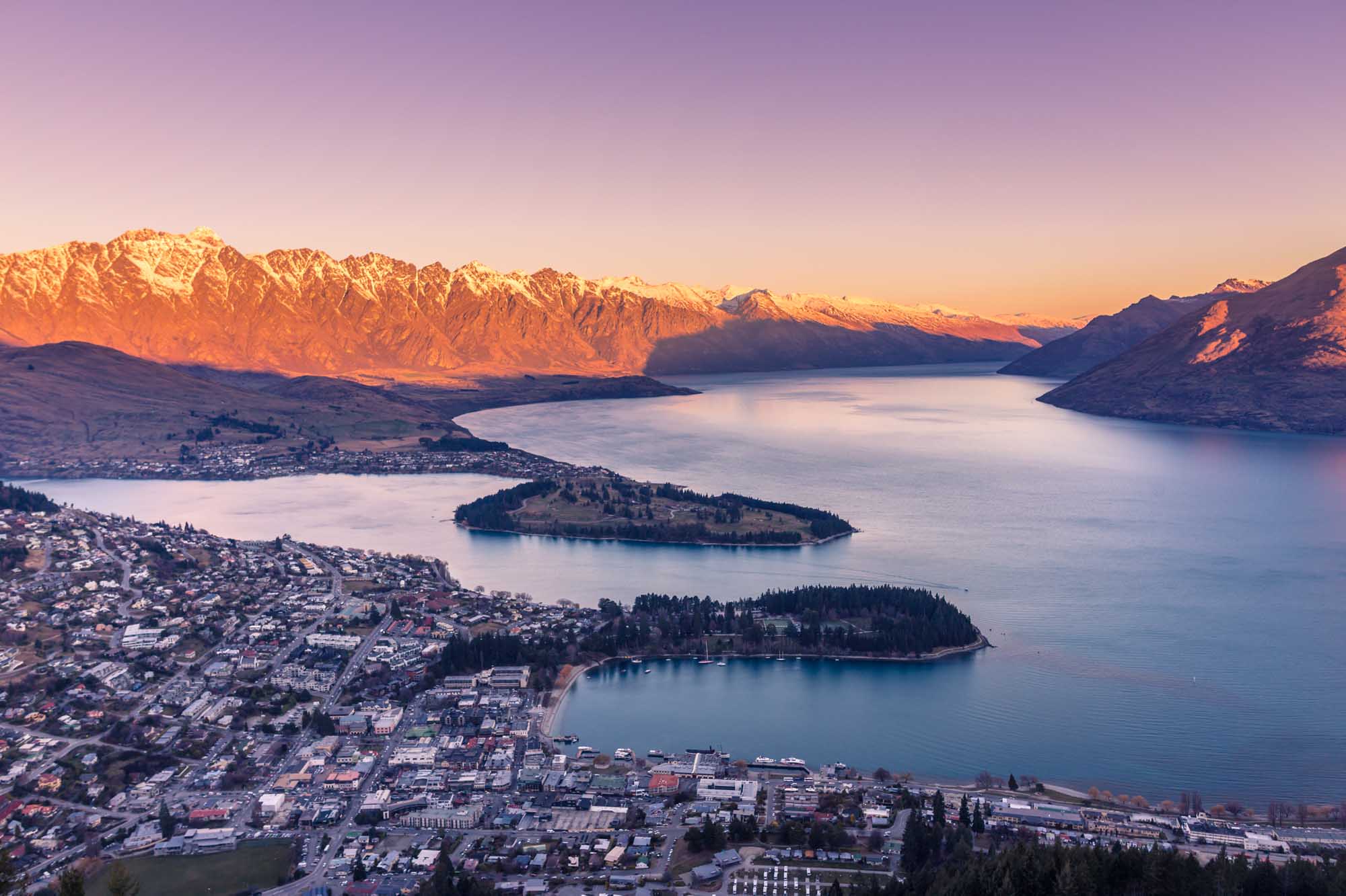 An aerial view of Queenstown, New Zealand at golden hour, showcasing the picturesque lakeside town