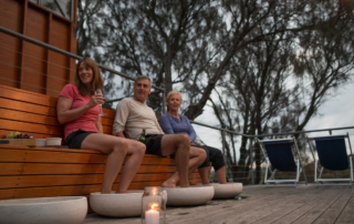 Three adults enjoy a leisurely afternoon on a wooden deck overlooking bare trees and water