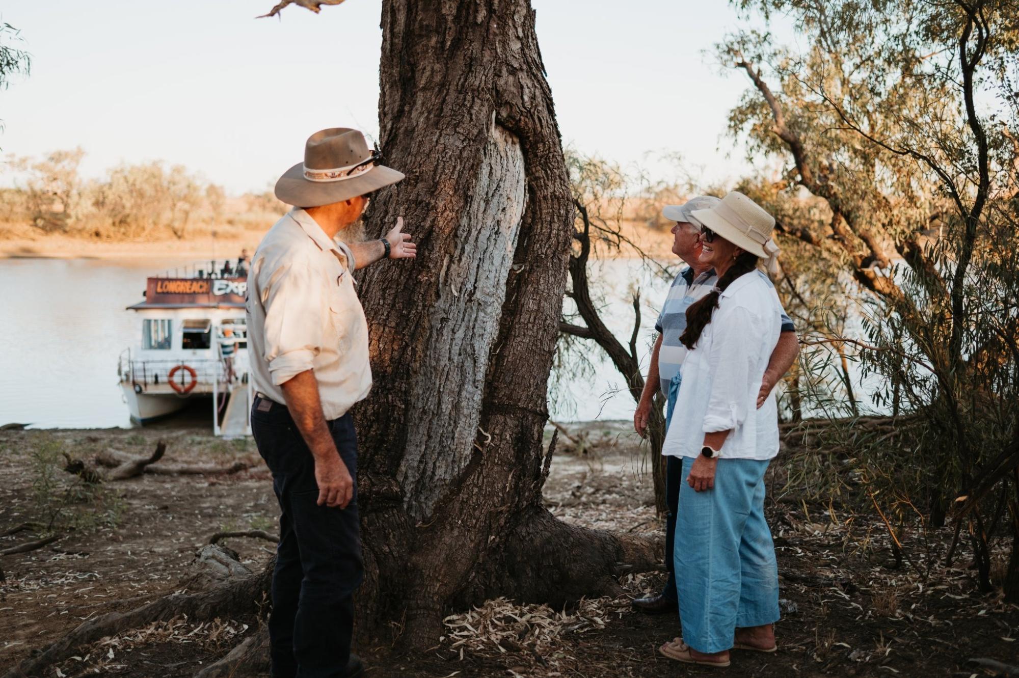 A uniformed crew member and two local guides stand beside a massive ancient tree on the riverbank