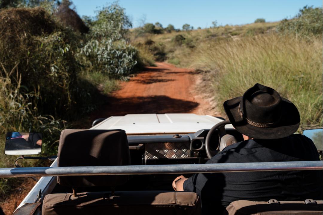 A person in a black cowboy hat drives an open-air safari vehicle along a distinctive red earth