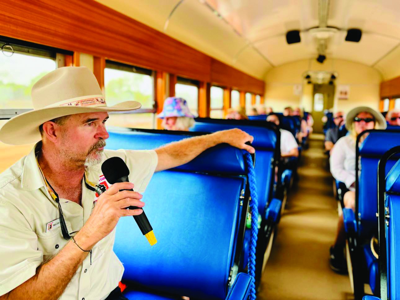 A tour guide wearing a cream-colored cowboy hat and light-colored uniform speaks into a microphone