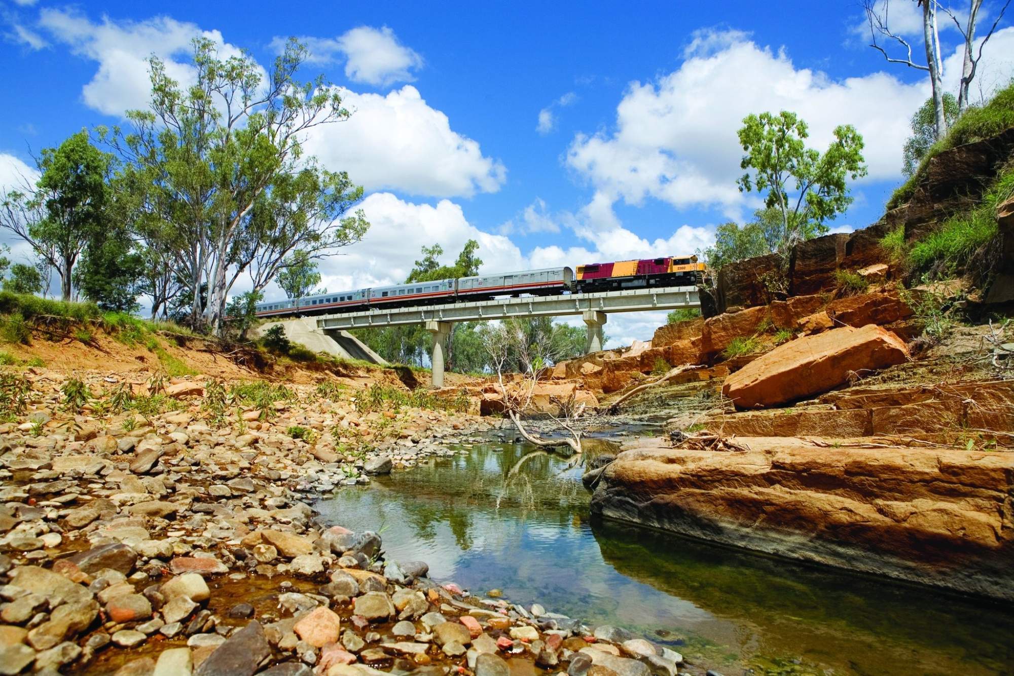 A scenic freight train with colorful locomotive crosses a modern bridge over a rocky creek bed