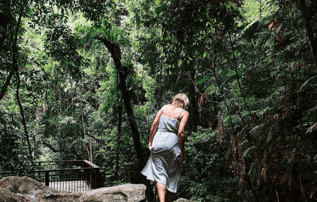 A woman in a light blue striped dress walks along a forested trail surrounded by dense, verdant