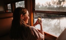 Woman Photographing Winter Landscape from Train Window A woman with long reddish hair sits by a train window, holding up a camera or mobile device