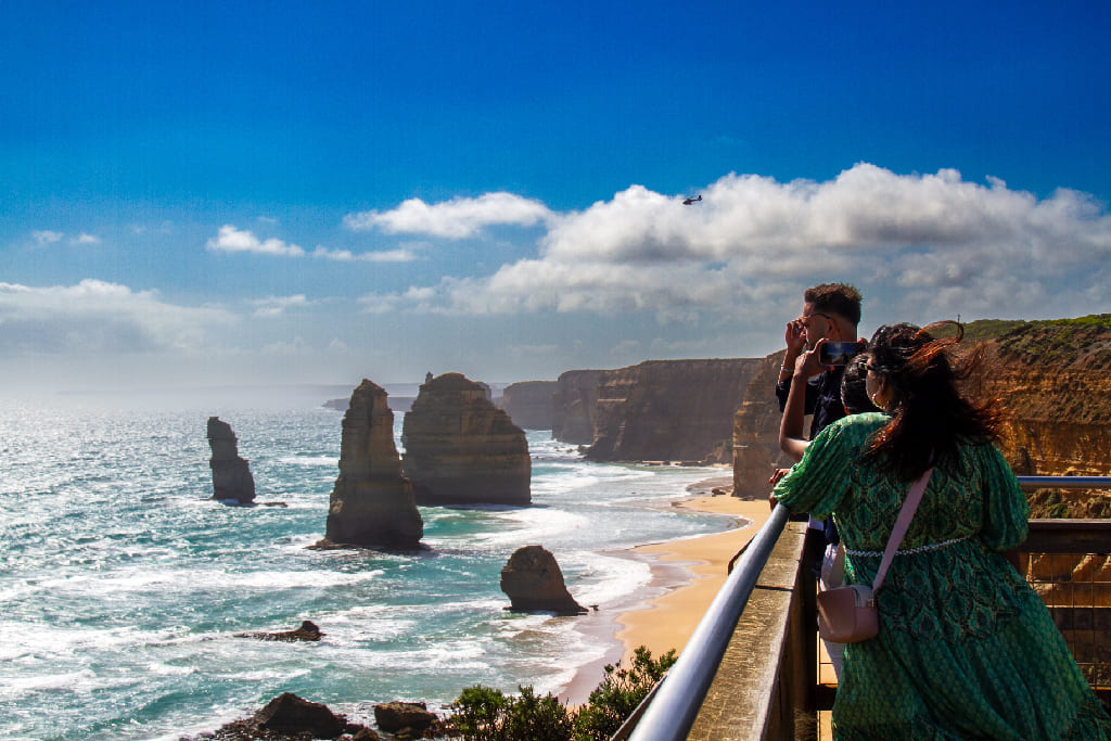 Visitors on a viewing platform look out over dramatic limestone sea stacks rising from the ocean beside sandstone cliffs, with a helicopter flying above.