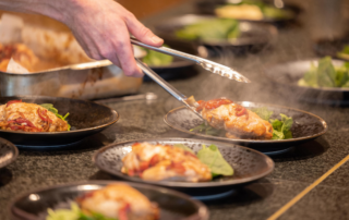 A server uses tongs to plate an elegantly presented dish featuring seared protein with red sauce