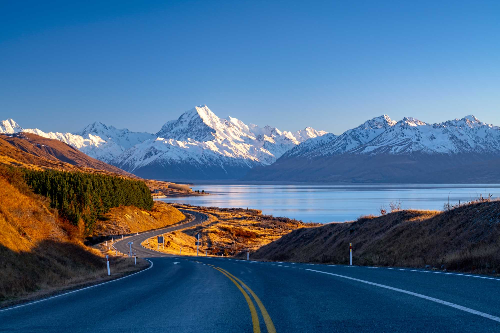 A scenic mountain road winds through golden tussock grassland toward snow-capped peaks reflected