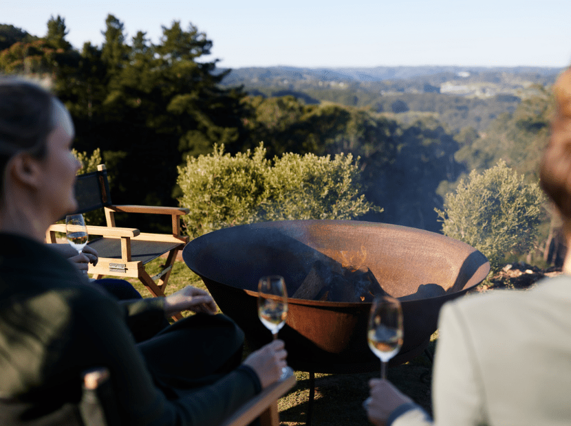 A group of people enjoy an alfresco meal at a table with wine glasses, overlooking a lush forested